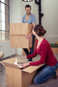 Happy young couple moving into a new home smiling at each other as the wife unpacks a carton on the floor as the husband arrives with two more.jpeg