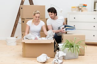 Happy young couple unpacking in their new home kneeling on a bare wooden floor unwrapping items from a large brown cardboard box.jpeg